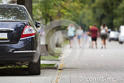 Back view part of black luxury shiny car parked on city pedestrian zone pavement on background of blurred silhouettes of people wa Stock Photo