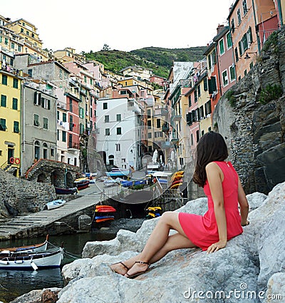Back view of red dressed girl sitting on the stones like a mermaid looking landscape of Italian Riviera, Riomaggiore, Cinque Terre Stock Photo