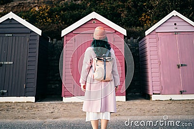 Back view stylish hipster woman with color hair in total pink outfit and backpack looking at wooden beach huts. Off Stock Photo
