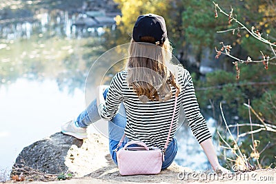 Back view on traveler hipster girl wearing black blazer. Young brave woman sittingon the edge of a cliff alone among trees in Stock Photo