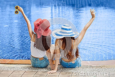 Back view of two beautiful Young Asian women in big summer hat and sunglasses sitting on the edge of the swimming pool with feet Stock Photo