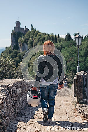 Back view of walking woman, dressed in a motorcycle outfit. body protection turtle and knee pads, helmet in hand. Fortress, stone Stock Photo