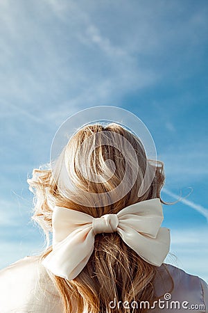 Back view of woman with long wavy fair hair decorated with beige bow made of ribbon standing on blue sky background. Stock Photo