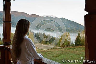 Back view of woman in spa bathrobe standing at wooden balcony of hotel resort room and enjoying beautiful morning alpain Stock Photo