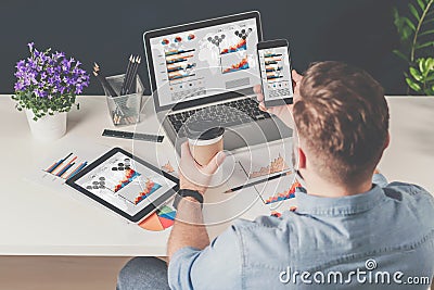 Back view.Young bearded businessman in denim shirt sits in office at table and uses smartphone with charts,diagrams Stock Photo