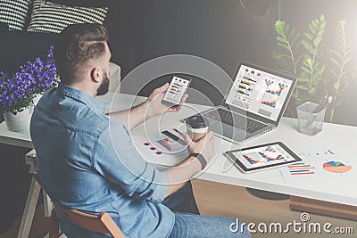 Back view.Young bearded businessman in shirt sits in office at table and uses smartphone with charts,diagrams and graphs Stock Photo