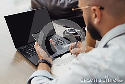 Back view. Young bearded man trendy glasses sits cafe front of laptop computer, uses smartphone, takes notes in notebook Stock Photo