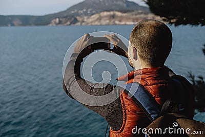 Back view of Young Male Traveler doing photo by phone on the background of the sea Stock Photo