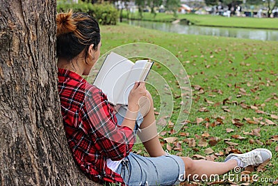 Back view of young relaxed man in red shirt leaning against a tree and reading textbook in beautiful outdoor park. Stock Photo