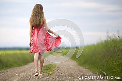 Back view of young romantic slim woman in red dress with long hair walking by ground road along green field on sunny summer day on Stock Photo