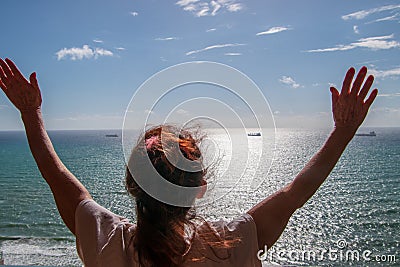 Back of woman with long red hair looking out at a calm flat ocean with her arms up in the air. Stock Photo