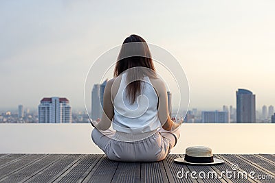 Back of woman relaxingly practicing meditation at the swimming pool rooftop with the view of urban skyline building to attain Stock Photo