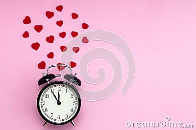 Black alarm clock and red hearts around it on a pink background. Place for text. Valentine`s Day. Time for love Stock Photo