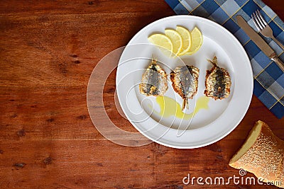 Background of a wooden table top with a plate of baked sardines on it, with bread, cutlery and a napkin on the edge, with space Stock Photo