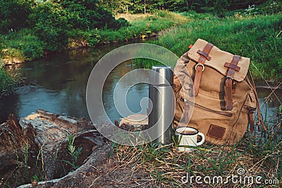 Backpack of traveller, thermos and Enameled mug of coffee or tea on bank of beautiful forest river Stock Photo