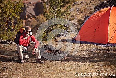 Backpacker having rest near orange tent, drinks tea from cup . Stock Photo