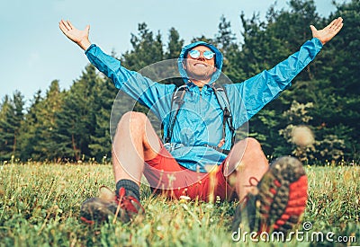 Backpacker man has a rest break enjoying mountain landscape wide opened and raised arms. He wears in blue rain coat poncho and Stock Photo