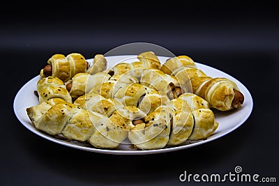 Baked ausages in the dough on a white plate against black background. Sausage Puff Pastry Buns.sausages wrapped in puff pastry and Stock Photo