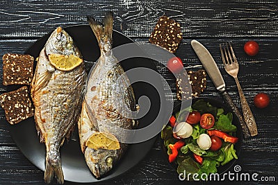 Baked Dorado fish on a black plate, salad with tomato mozzarella and lettuce leaves. Stock Photo