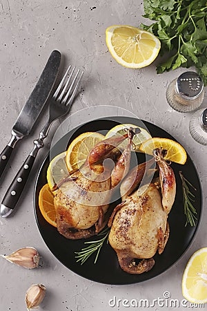 Baked quails with lemon and orange served on a dark plate on a gray background, Top view, Vertical orientation Stock Photo