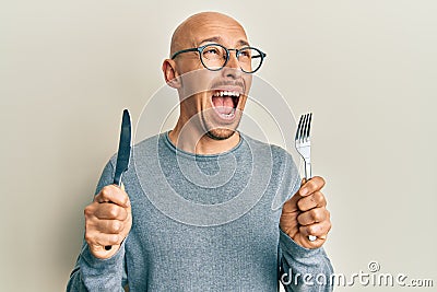 Bald man with beard holding fork and knife ready to eat angry and mad screaming frustrated and furious, shouting with anger Stock Photo