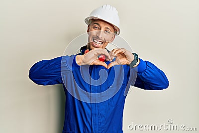 Bald man with beard wearing builder jumpsuit uniform and hardhat smiling in love showing heart symbol and shape with hands Stock Photo