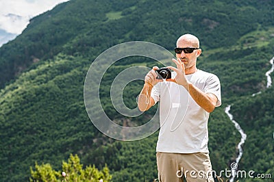 A bald tourist in sunglasses at a halt takes a picture on his camera. Against the backdrop of green mountains and a Stock Photo