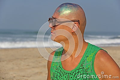 A bald and unusual young man, a freak, with a shiny bald head and round wooden glasses on the background of the beach and the sea Stock Photo