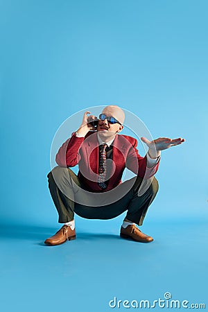 Balded man with mustache wear in vintage style squatting and talking on retro phone looking away over blue background. Stock Photo