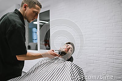 Barber with hair clipper in the hands makes a hairstyle in the beard of the client, lying on a chair and looking away with his Stock Photo