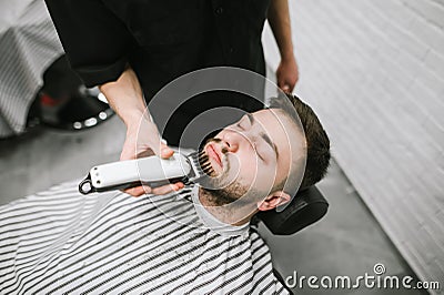 Barber with hair clipper in the hands makes a hairstyle in the beard of the client, lying on a chair. Male hairdresser doing Stock Photo