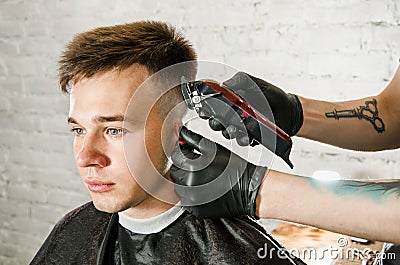 Barber hand in gloves cut hair and shaves young man on a brick wall background. Close up portrait of a guy Stock Photo
