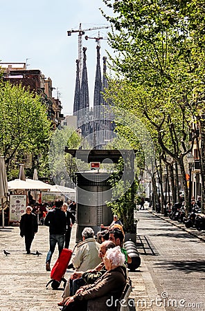 Barcelona city street scene, people relaxing on the benches. BasÃ­lica de la Sagrada Familia by Antoni Gaudi in the distance Editorial Stock Photo