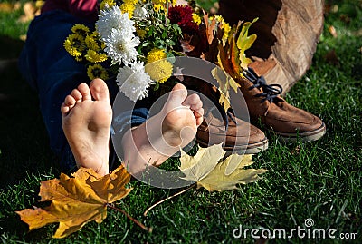 bare feet of child lying on grass next to a bouquet of autumn leaves in brown shoes on a warm sunny autumn day Stock Photo