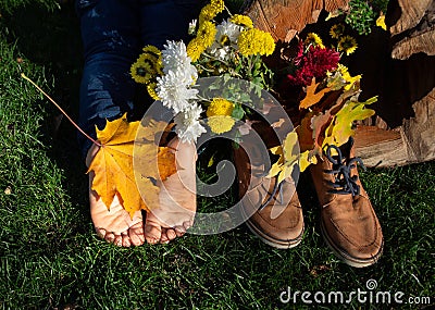 bare feet of child lying on grass, next to brown shoes, bouquet of chrysanthemums and leaves on sunny autumn day Stock Photo