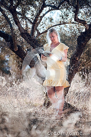 A barefoot blonde woman in a yellow summer dress is reading a book in the shade of olive trees on a beautiful sunny day Stock Photo