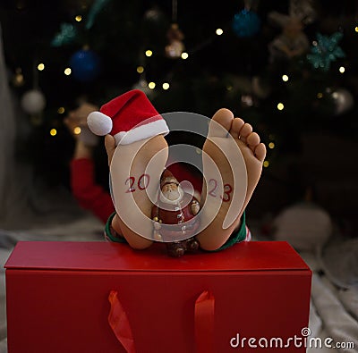 barefoot kid lies in front of the Christmas tree, the numbers 2023 are written on his feet, a souvenir of Santa Stock Photo
