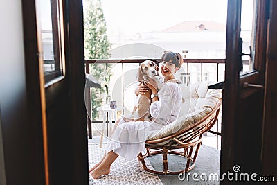 Barefooted laughing girl looking at camera, holding beagle dog while sitting in cozy chair. Lovely young lady with brown Stock Photo