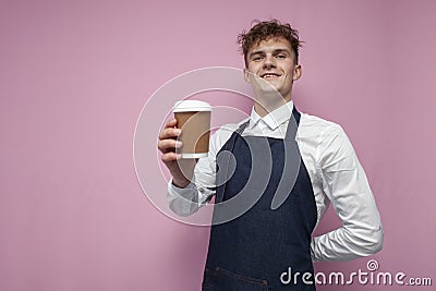 Barista in a white shirt and apron holds a cup of coffee and smiles on pink background, portrait of waiter worker guy Stock Photo