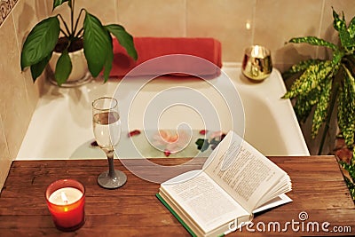 Bath with flower petals. Book, candles and glass of wine on a wood tray. Organic Spa Relaxation in comfort cozy bathroom Stock Photo