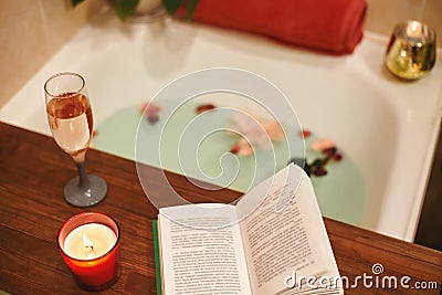 Bath with flower petals. Book, candles and glass of wine on a wood tray. Organic Spa Relaxation in comfort cozy bathroom Stock Photo