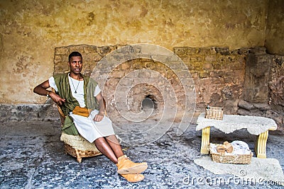 Black reenactor in costume poses as Roman scholar inside stone room with wooden book and worktable inside Editorial Stock Photo