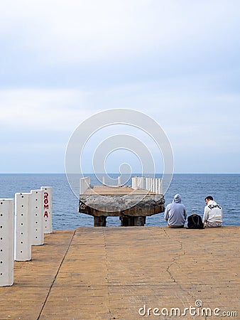 Two sit on the pier with their legs dangling. Two guys at sea. Friends met and talk on the old pier. Destroyed infrastructure at Editorial Stock Photo