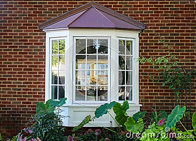 Bay window in a brick house with reflection of trees and view of windows and flowers inside and flowers and elephant ears outside Stock Photo