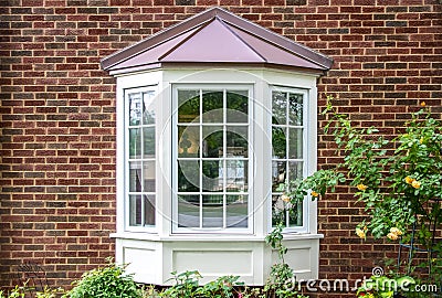 Bay window with copper roof on traditional brick home with flowers below and yellow roses extending out onto the window in Editorial Stock Photo