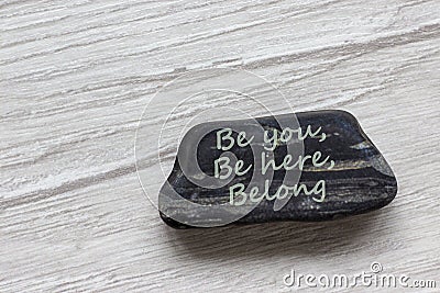 Be here, be you, belong symbol. Beautiful stone with words `Be here, be you, belong` on beautiful white wooden background. Stock Photo