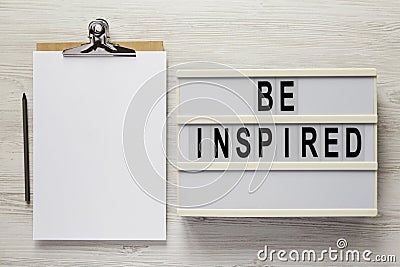 `Be inspired` on a lightbox, cliboard with blank sheet of paper on a white wooden background, top view. Flat lay, overhead, from Stock Photo