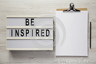 `Be inspired` on a lightbox, cliboard with blank sheet of paper on a white wooden surface, top view. Flat lay, overhead, from Stock Photo