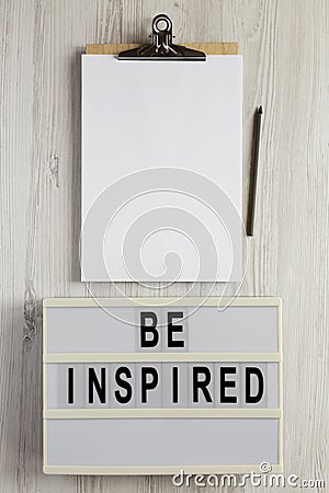 `Be inspired` on a lightbox, cliboard with blank sheet of paper on a white wooden surface, top view. Flat lay, overhead, from Stock Photo