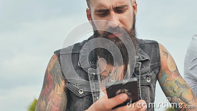 Bearded and brutal man with tattoos looks into the phone on the street. Stock Photo
